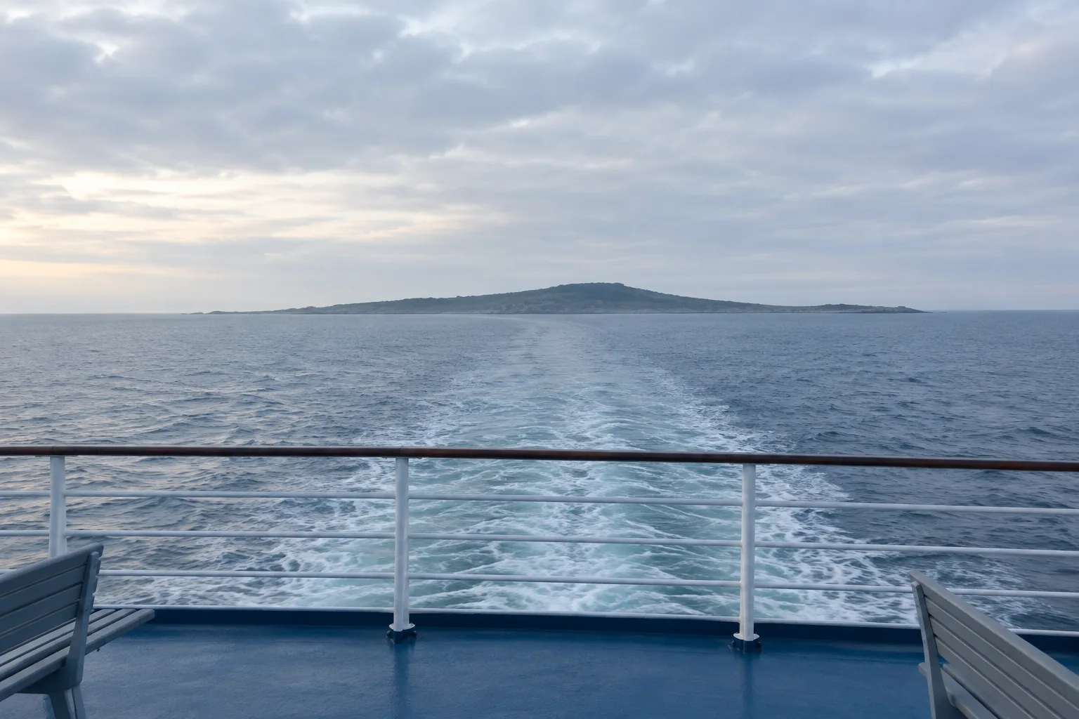 Depuis le pont d’un ferry, une rambarde encadre le sillage et la côte lointaine.