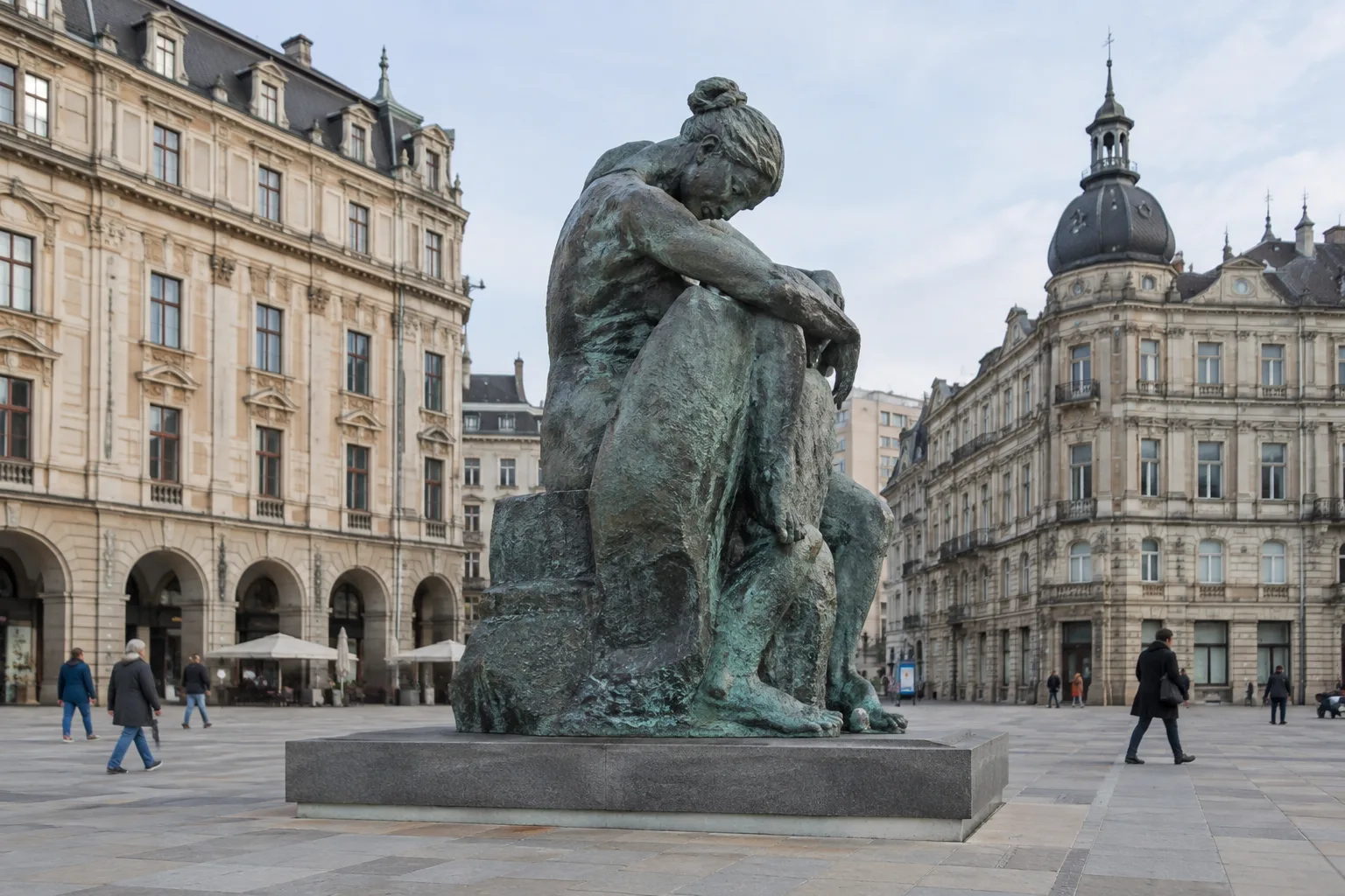 Des passants éloignés traversent une place devant une sculpture, dont l’échelle se mesure au décor.
