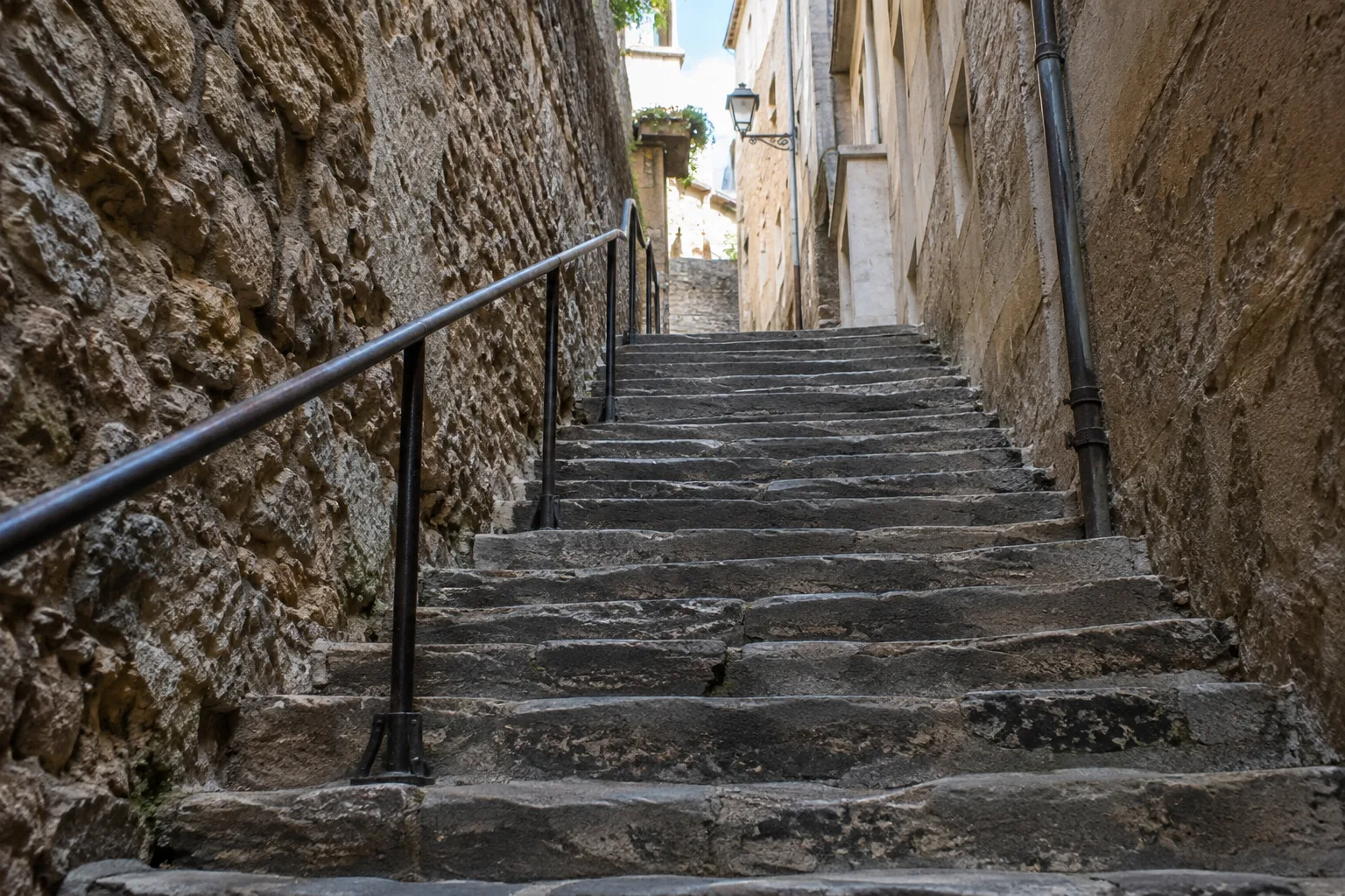 Un escalier de pierre ancien vu depuis le bas, dont les marches et la rampe montent vers une ouverture lumineuse.