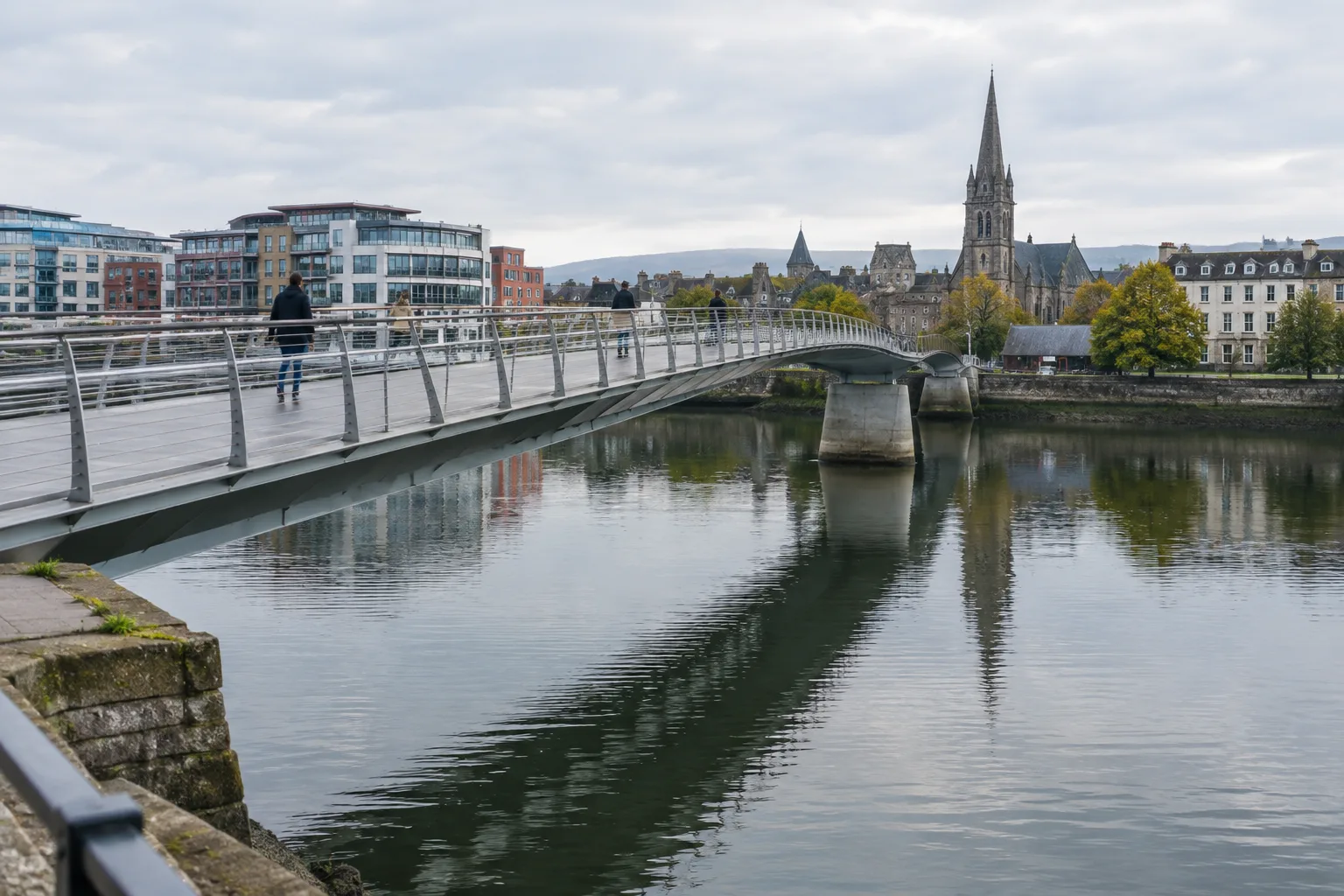 Un pont piétonnier traverse une rivière urbaine, avec quelques passants visibles au loin.