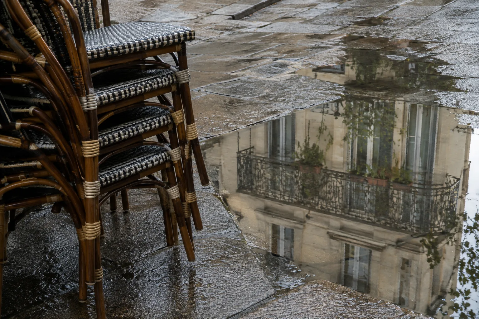 Des chaises empilées devant un café voisinent avec une flaque où se reflète un balcon.